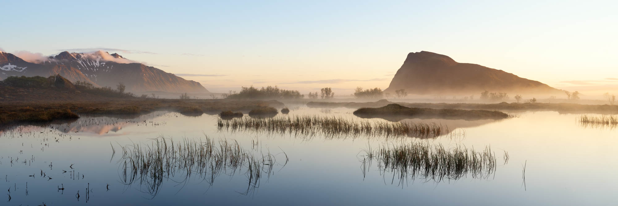 Panorama of a lake and Hoven mountain revealed in the mist in Gimsøymyrene nature reserve on Gimsøy Island in the Lofoten Islands during the midnight sun