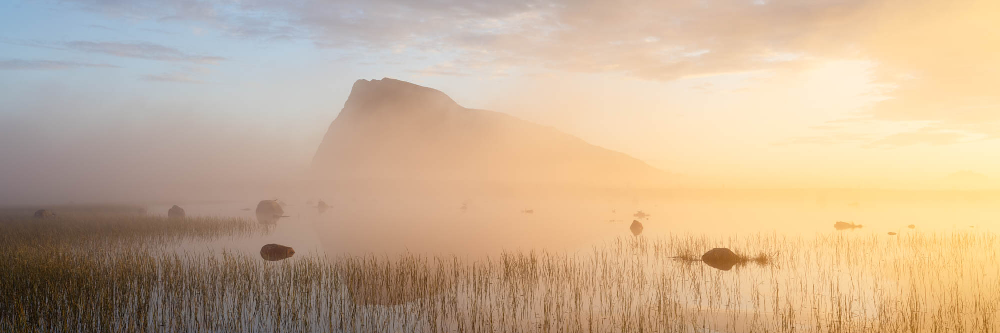 Panorama of a lake and Hoven mountain revealed in the mist in Gimsøymyrene nature reserve on Gimsøy Island in the Lofoten Islands during the midnight sun