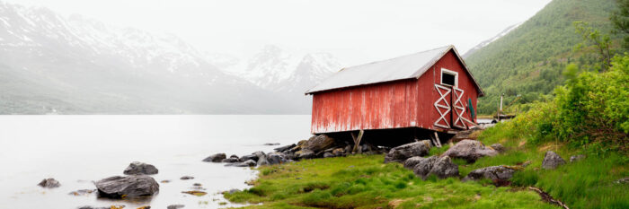 Panorama of an ageing and weathered wonky red Norwegian boathouse blown over in the Arctic winds