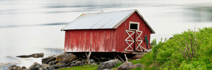 Panorama of an ageing and weathered wonky red Norwegian boathouse